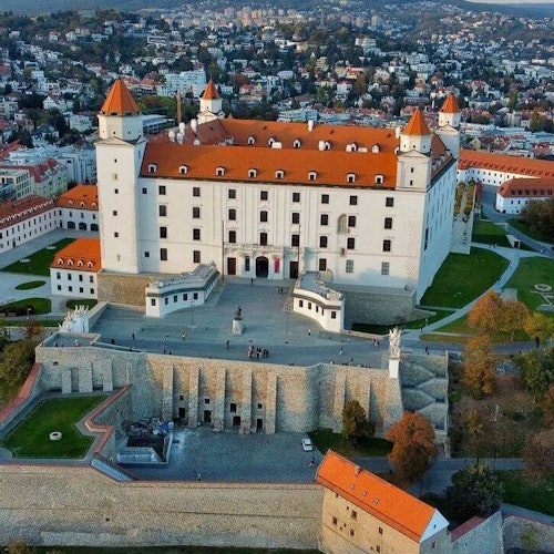 Vista aérea de un gran castillo blanco con tejados rojos, rodeado de vegetación y un pueblo al fondo.