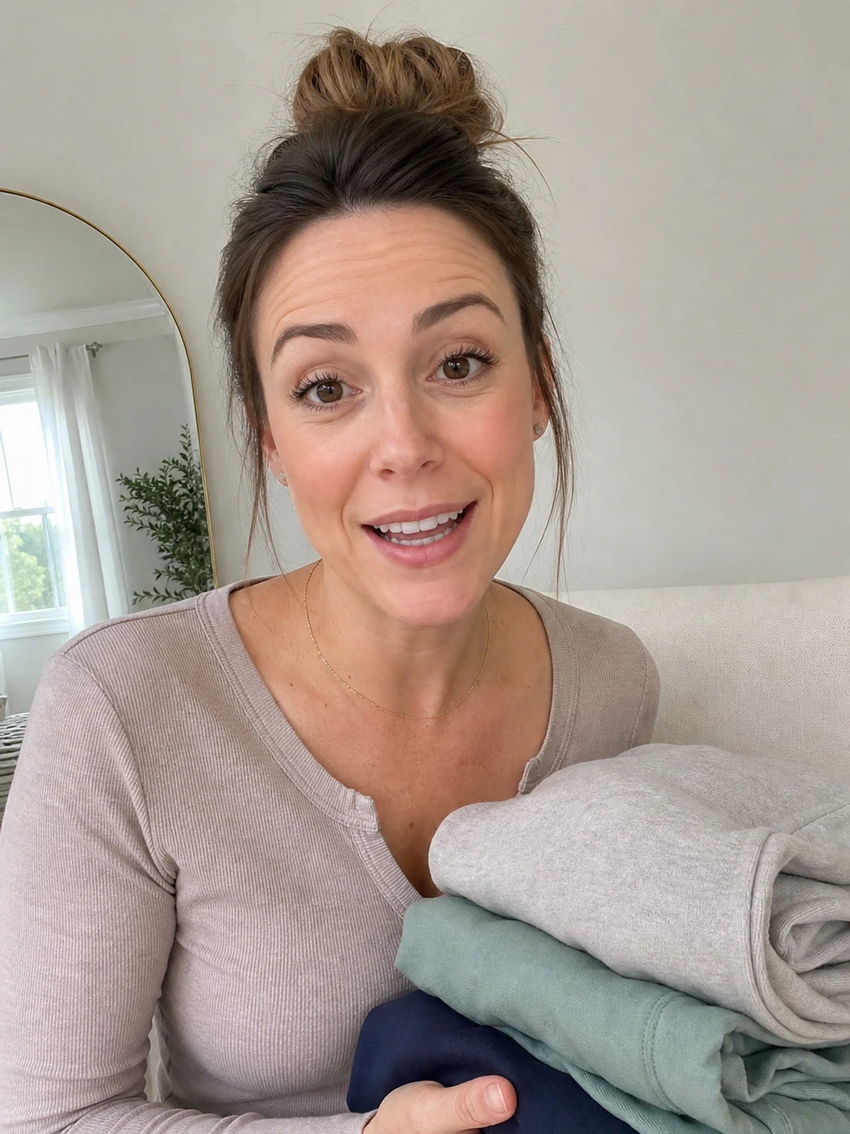 UGC-style photo: parent creator, in a bright home studio, holding folded apparel while talking to camera, mirror-style vertical shot.