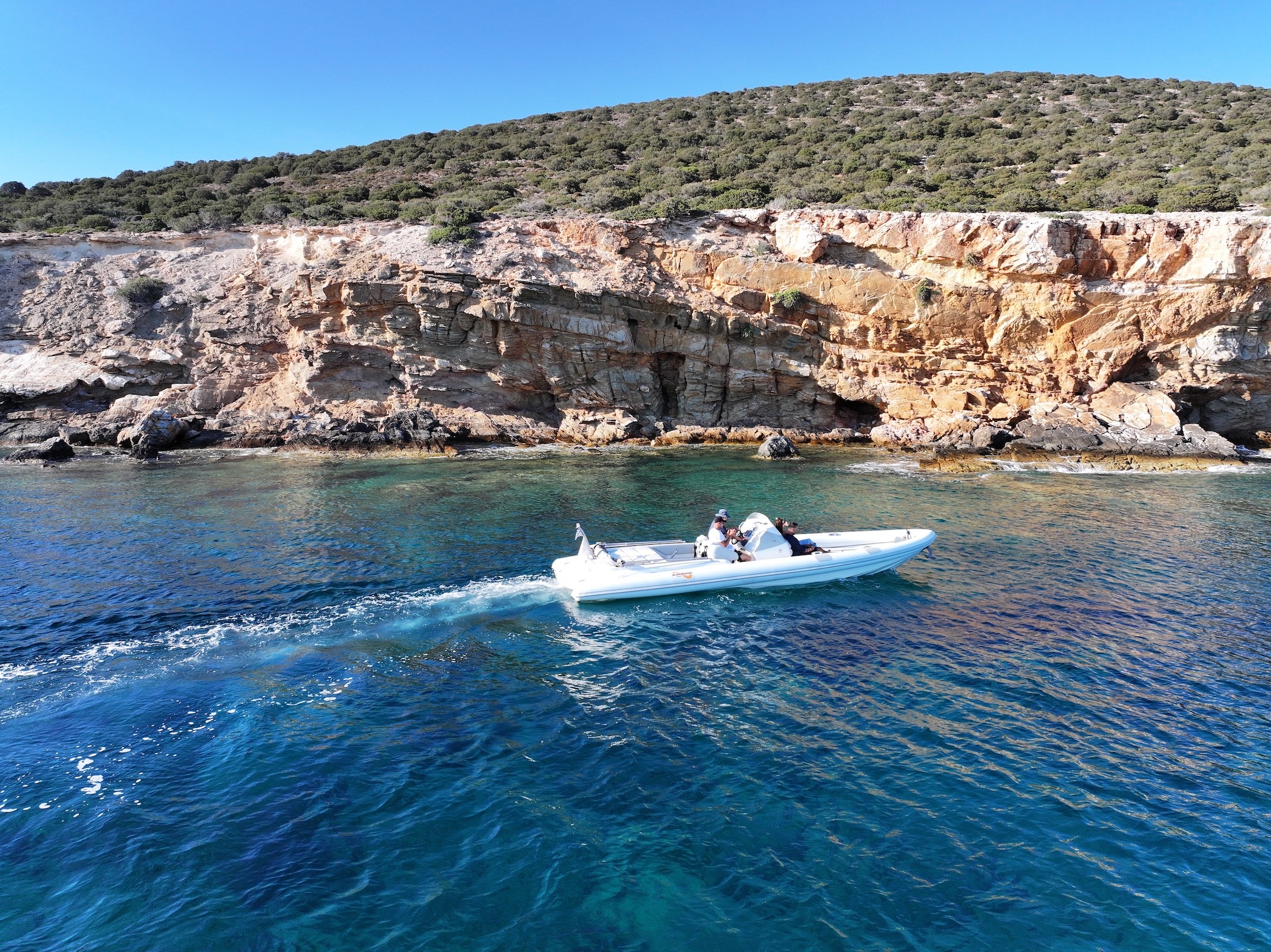 White motor yacht anchored in a secluded rocky cove with sparkling turquoise waters and dramatic limestone cliffs under bright Mediterranean sun.