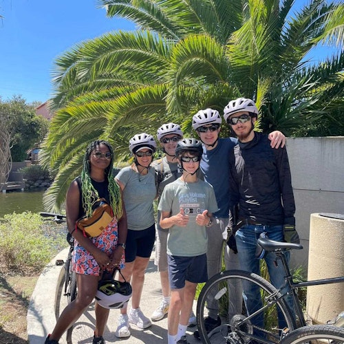 Un grupo de seis personas con cascos posa con sus bicicletas frente a palmeras en un día soleado.