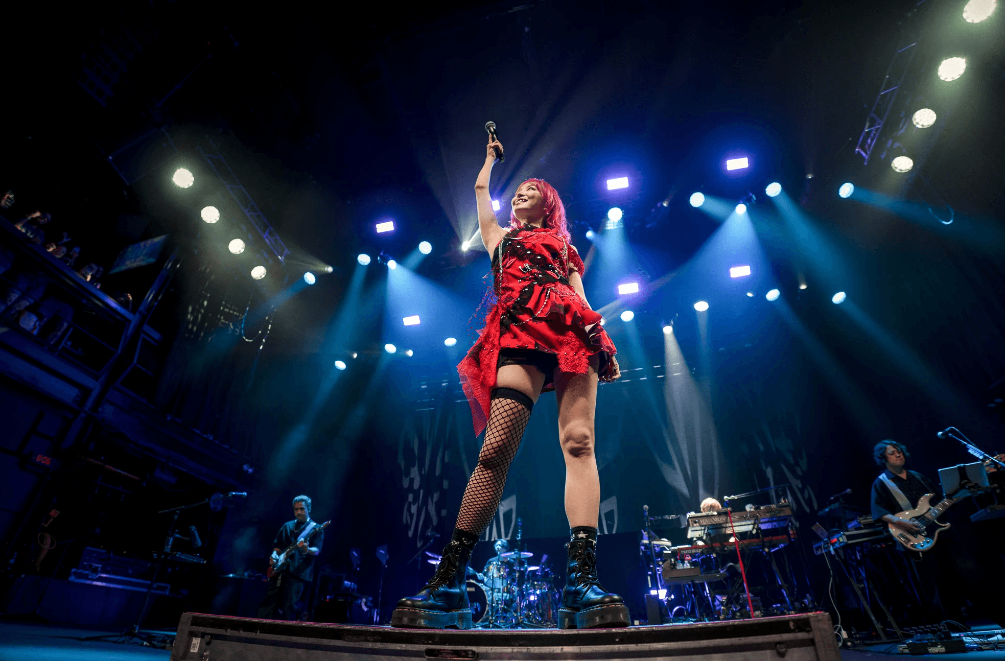 LISA A singer in a red dress and black boots performs energetically on stage, surrounded by bright stage lights and musicians playing instruments at a lively concert venue.