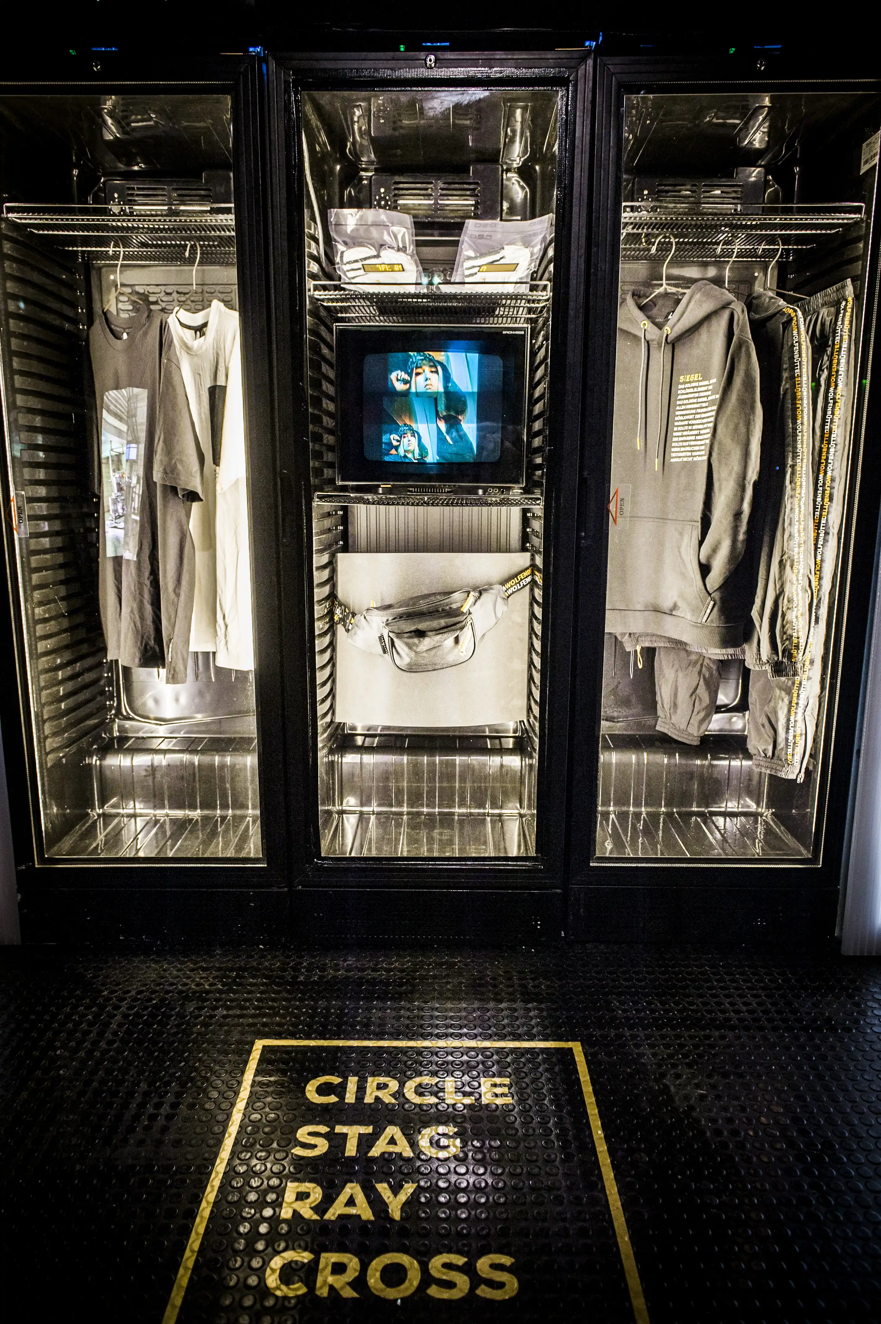 Streetwear pieces presented inside illuminated fridge cabinets with a tube TV screen at the JÄGERMEISTER launch, with yellow Circle Stag Ray Cross lettering on the black floor.