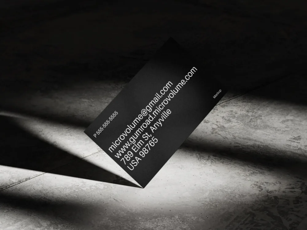 Minimalist black business card mockup displayed on a concrete surface. The card is propped up, featuring white contact details in a clean font. Dramatic lighting casts sharp shadows for a modern look.