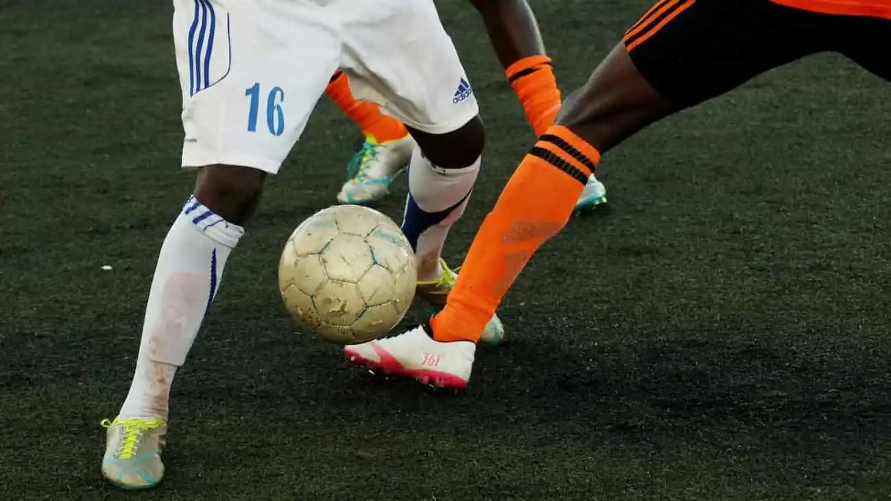 Three football players in an intense duel for the ball - two players in orange jerseys and one player in white on the green pitch, symbolising fair and performance-oriented competition in the context of a performance comparison tournament.