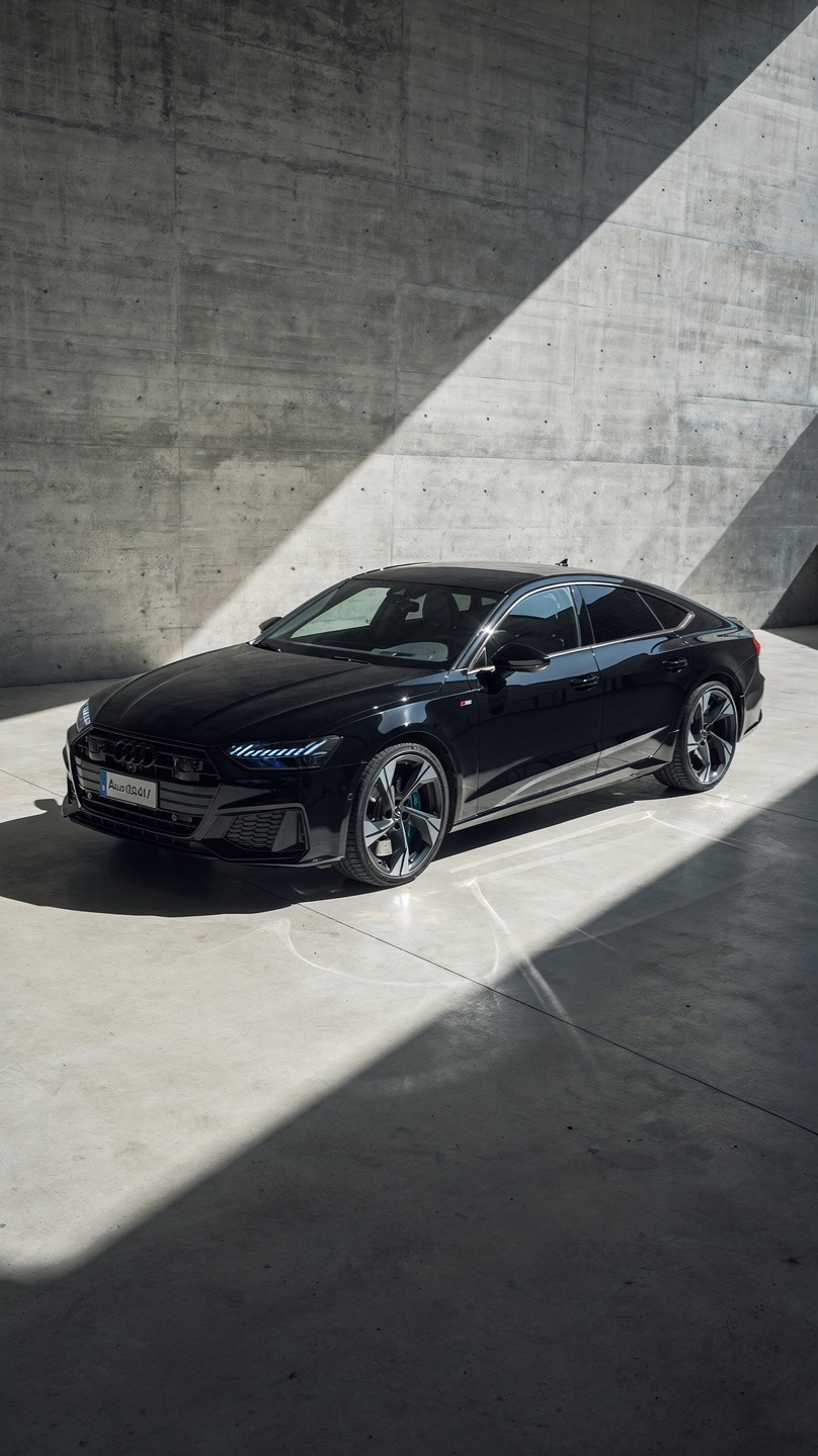 Sleek black car in a modern concrete space, partially lit by a diagonal beam of sunlight, creating a dramatic contrast. The scene feels sophisticated and elegant.