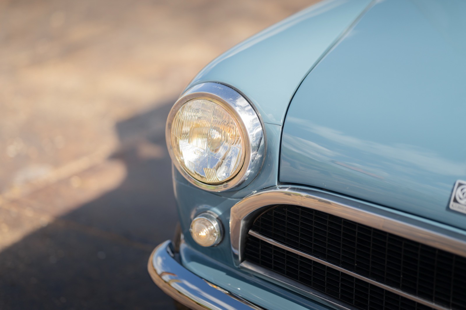 Close-up of a vintage car's headlight and chrome detailing, showcasing a soft blue color against a textured background.