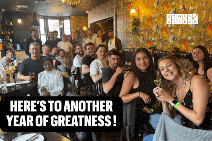 Group gathered in a restaurant with ornate yellow wallpaper. Text overlay at bottom reads 'HERE'S TO ANOTHER YEAR OF GREATNESS!' with StoryStream logo in top right corner.