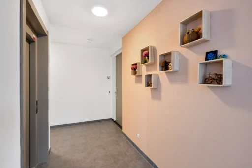 Een gang met een lichtroze wand en kleine wandkastjes met planten en decoratie.