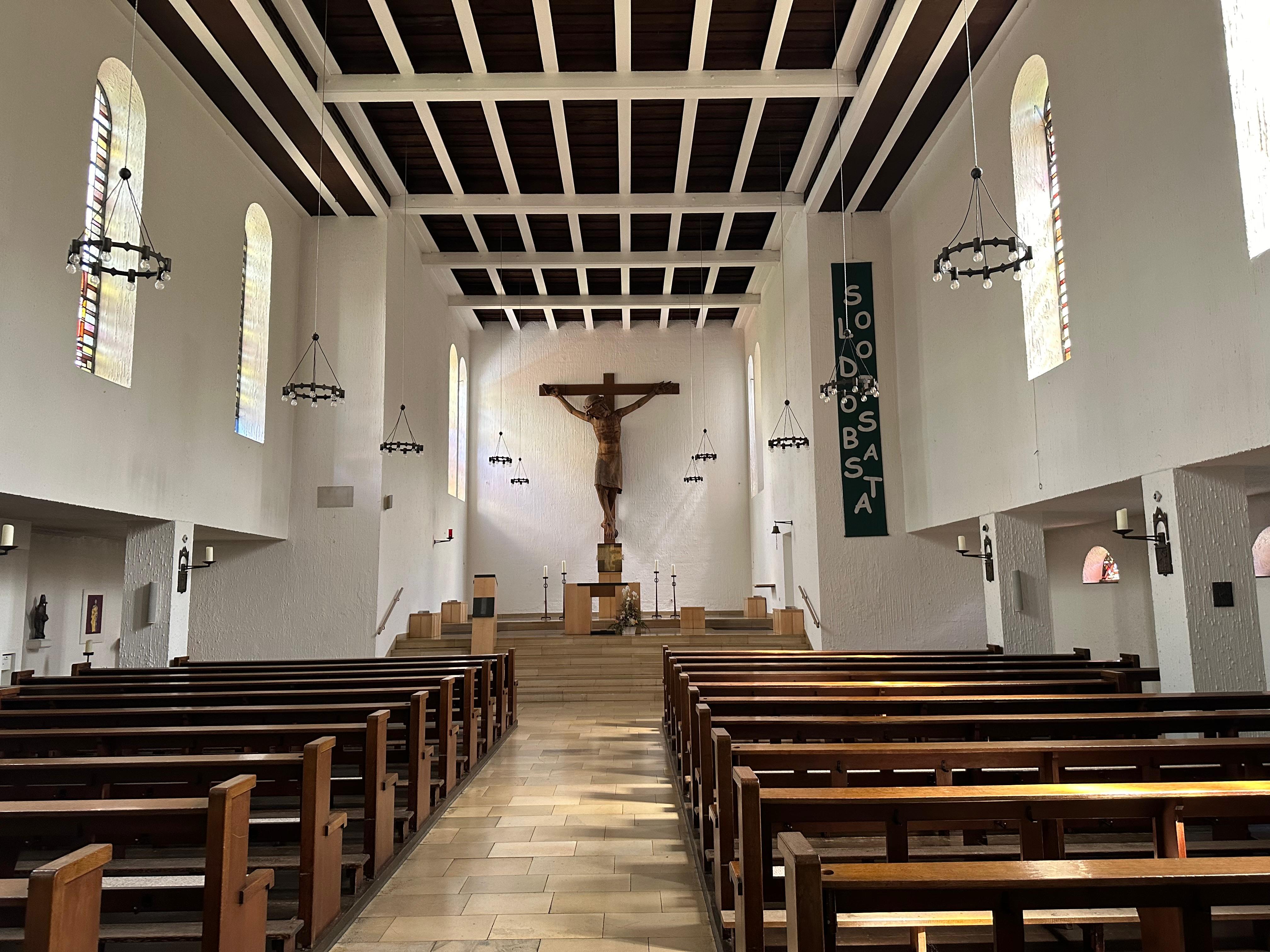 Saal der Marienkirche. EIn geräiumiger Saal mit Kirchenbänken, weißen Wänden und einem großen Kreuz mittig auf der Bühne.