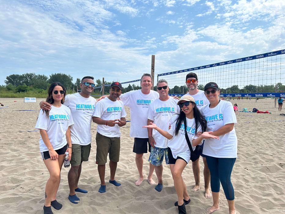 A large group of adults and children posing for a photo on a sandy beach in front of a giant blue inflatable "SickKids" sign during the Heatwave for SickKids event.