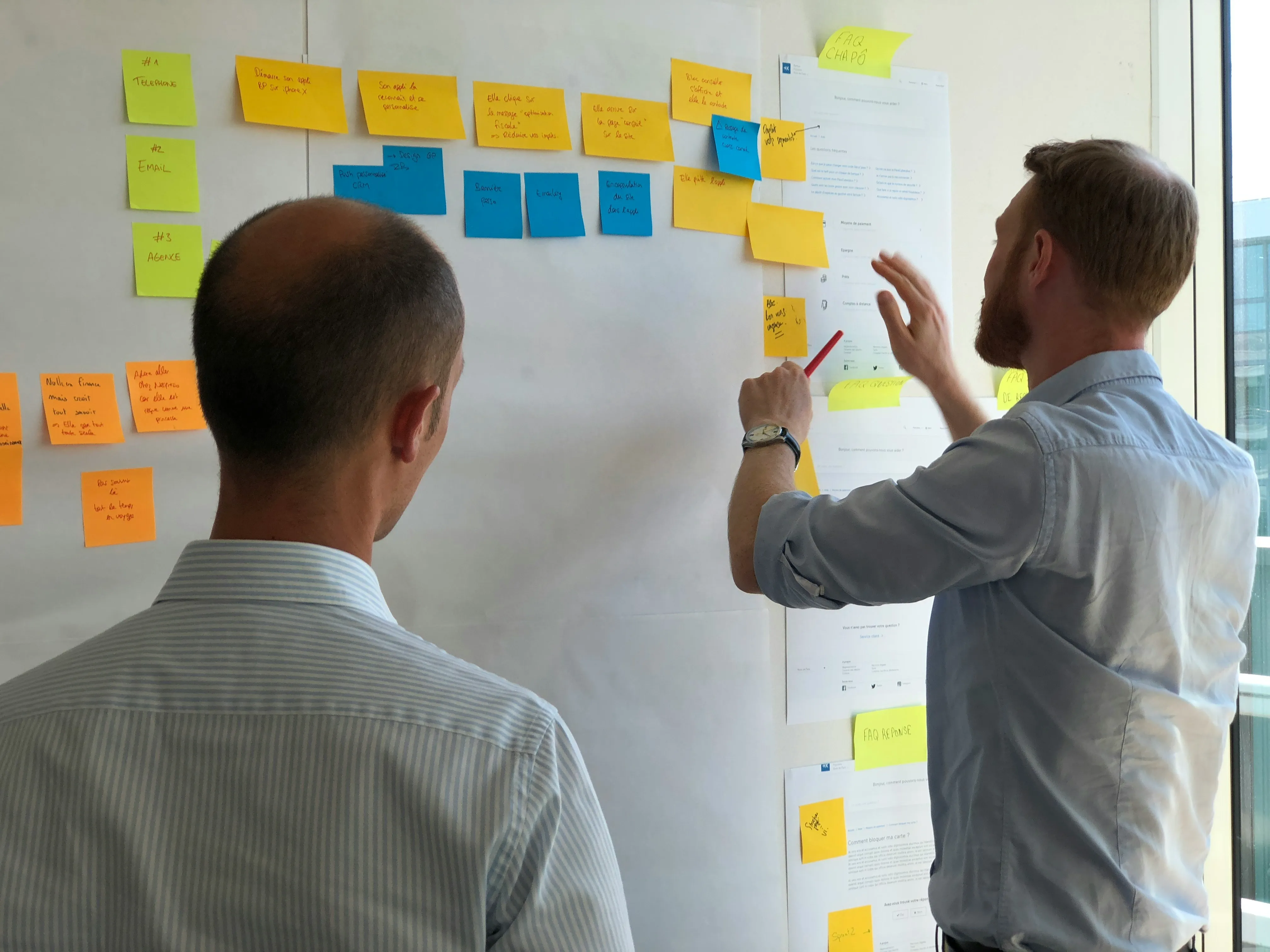 Two people discuss ideas using sticky notes on a wall in a bright, modern workspace.