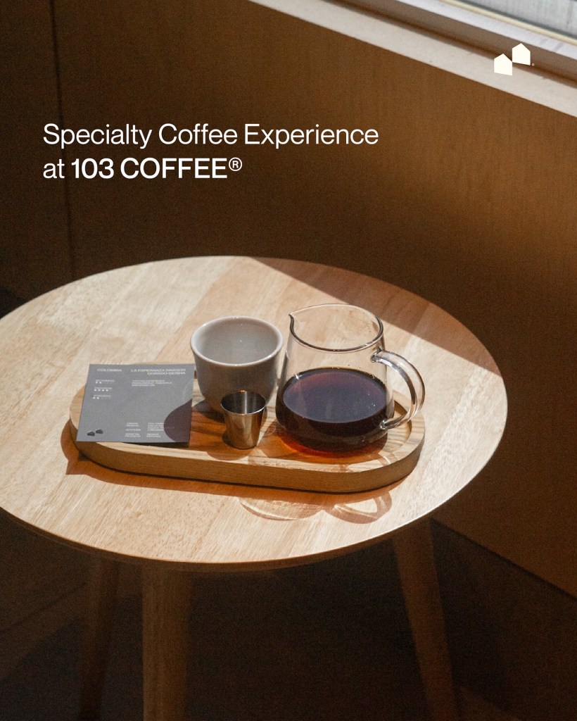 A wooden table featuring a specialty coffee experience at 103 Coffee, including a cup, a small jug, a shot glass, and a menu item on a wooden tray.