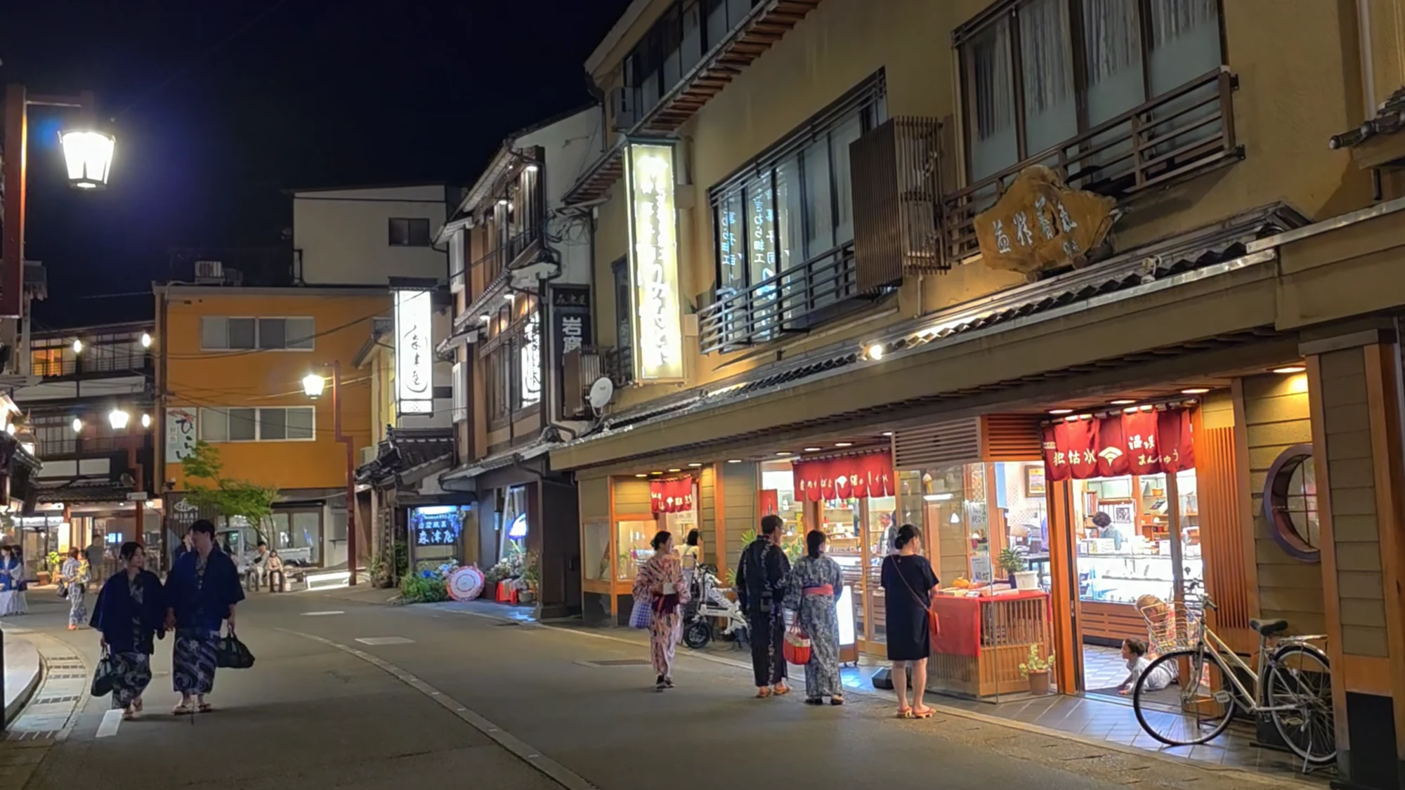 People in yukata walking a lit Kinosaki shopping street in the evening.