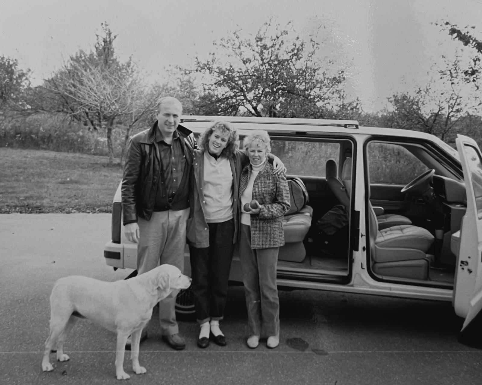 Black-and-white photo of three people, including Normy, standing arm-in-arm beside an open vehicle, with a dog in the foreground; an early moment in the journey of home textiles entrepreneurship.