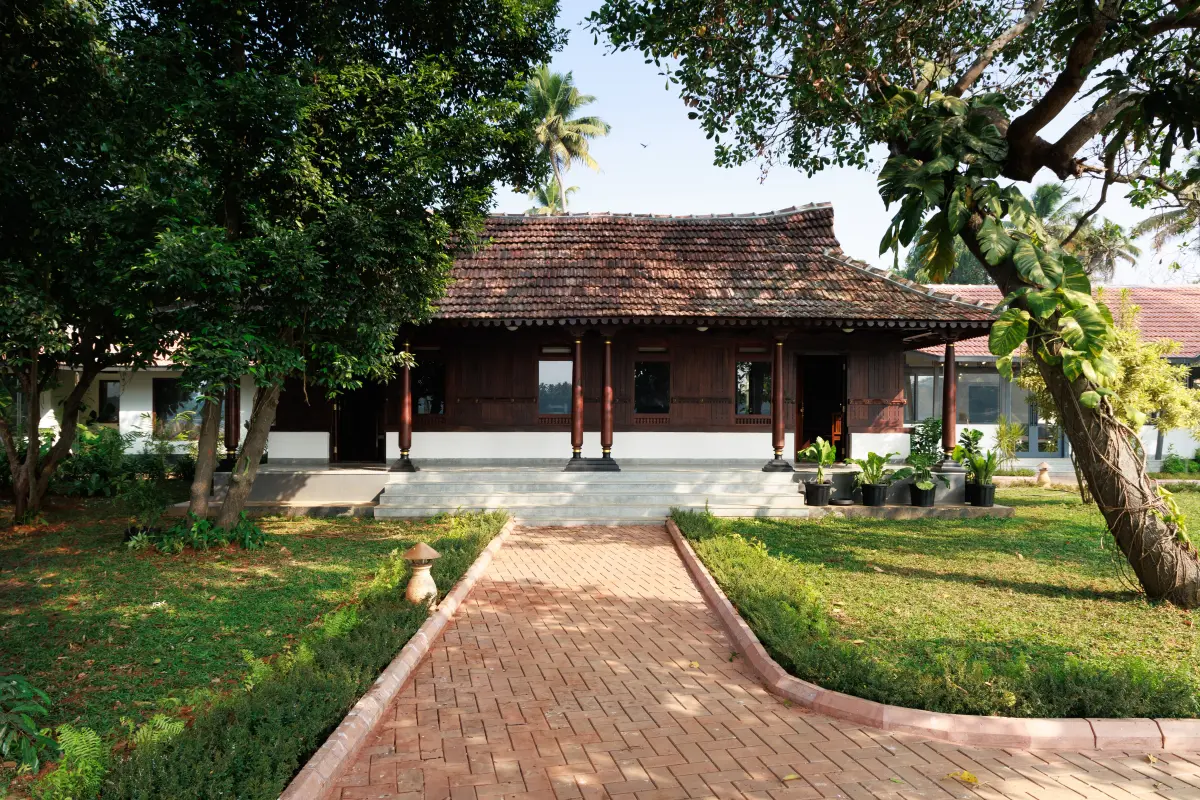 Traditional Kerala tharavadu entrance at Vedic Village Kodungallur heritage resort, with carved dark wood pillars, terracotta roof and brick pathway through tropical garden
