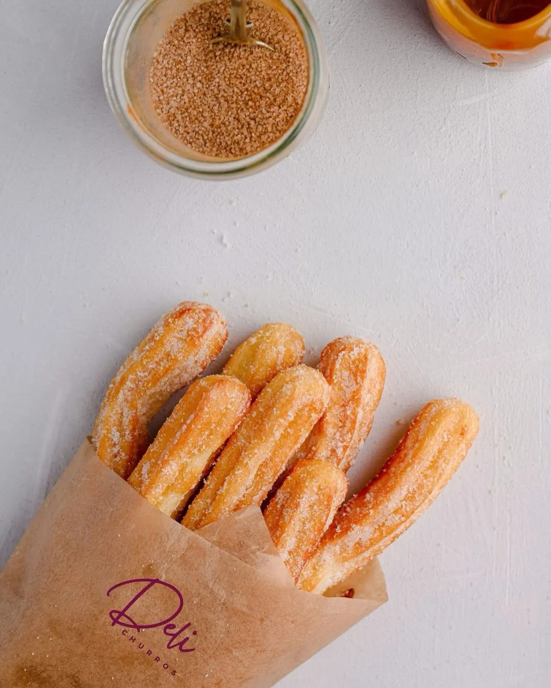 Sugar-dusted churros spilling from a Delichurros paper cone beside a jar of cinnamon sugar