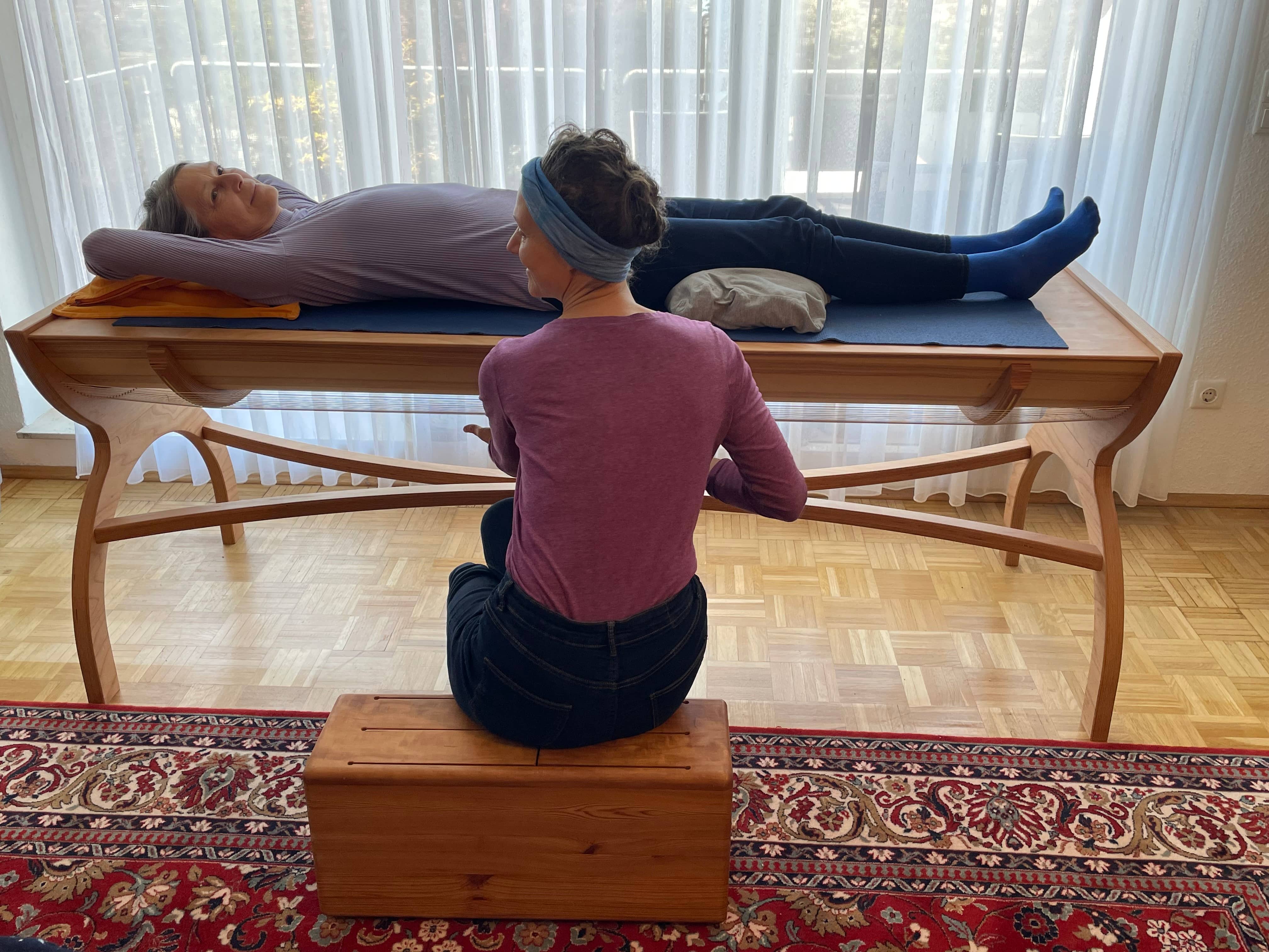 Therapeut leitet Klientin auf Massageliege an. Klientin liegt auf dem Rücken, Therapeut sitzt.