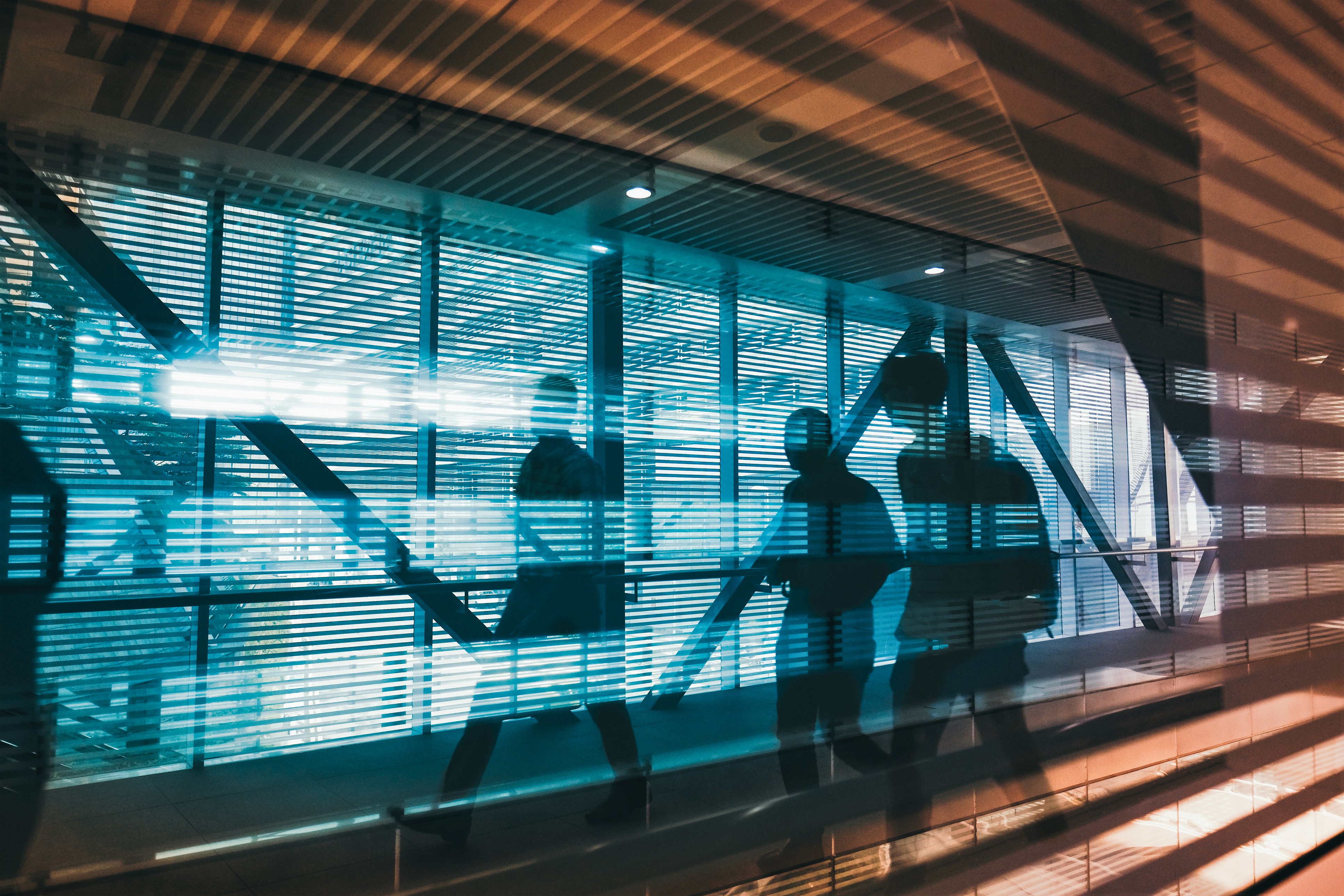 Silhouetted figures walking through a modern building hallway with glass windows and blinds reflecting light.
