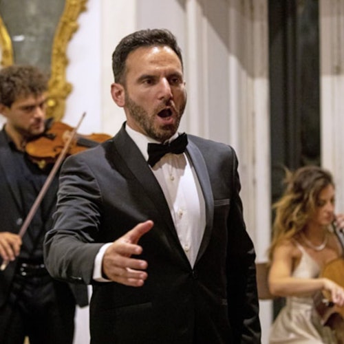 A man in a black tuxedo sings passionately, with a violinist and cellist performing in the background indoors.
