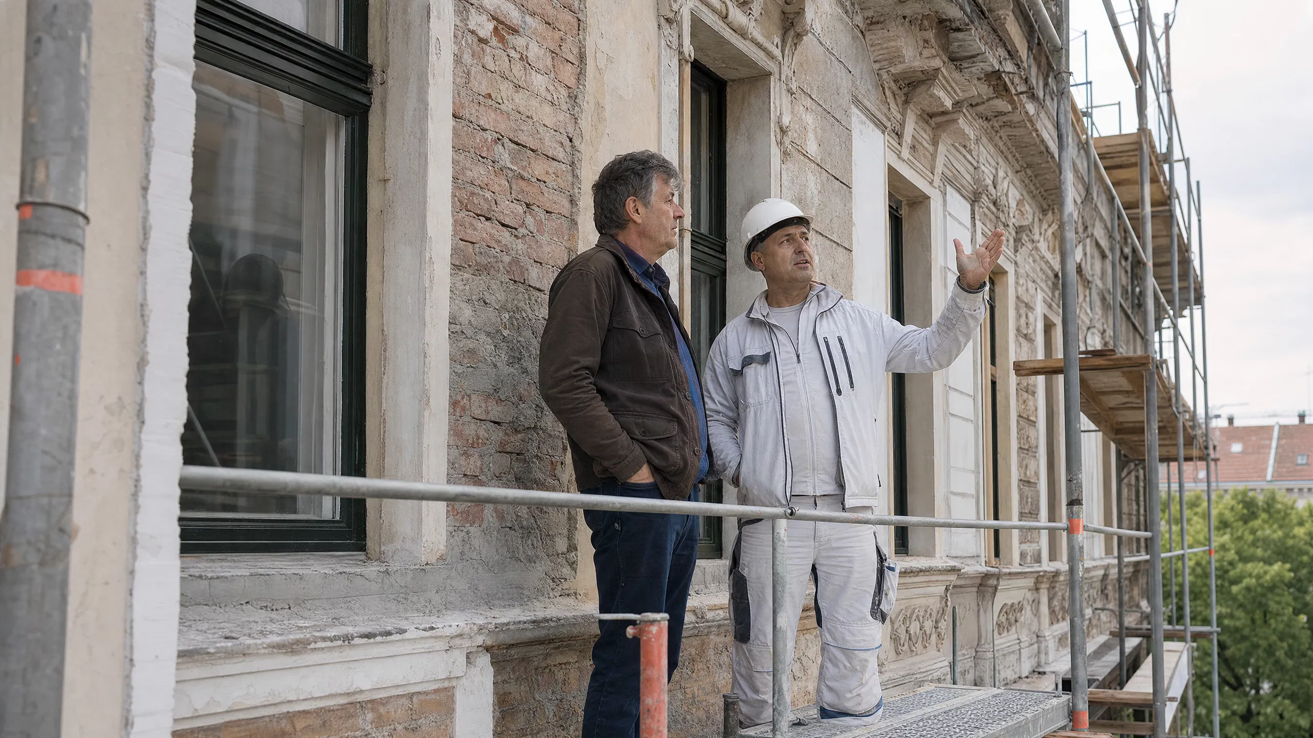 Ein Vermieter und ein Baupolier besprechen auf dem Gerüst eines gründerzeitlichen österreichischen Zinshauses den Zustand der teilweise freigelegten Fassade.
