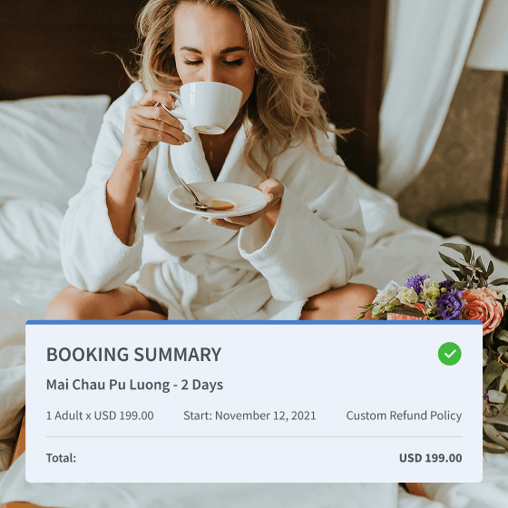 Woman in a white bathrobe sitting on a hotel bed, drinking coffee with a breakfast tray and bouquet of flowers beside her.