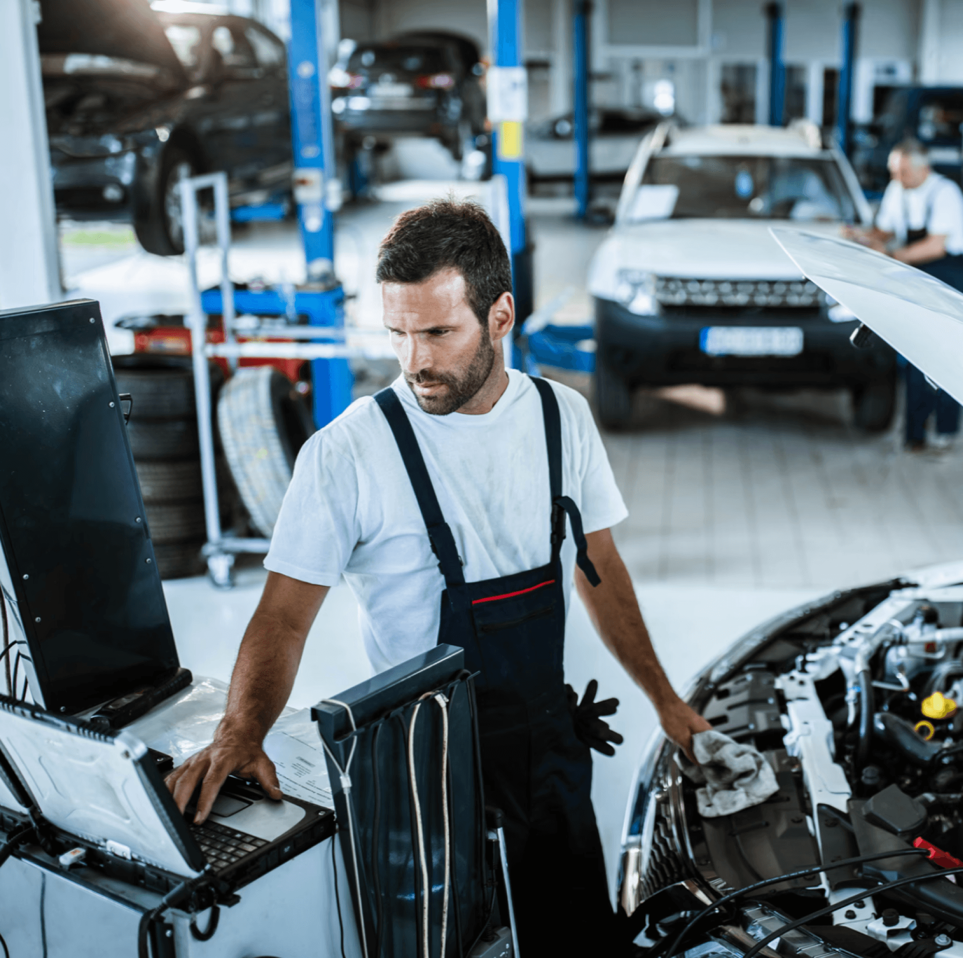 man in work clothes standing next to an open car hood and a computer inside of an auto repair shop