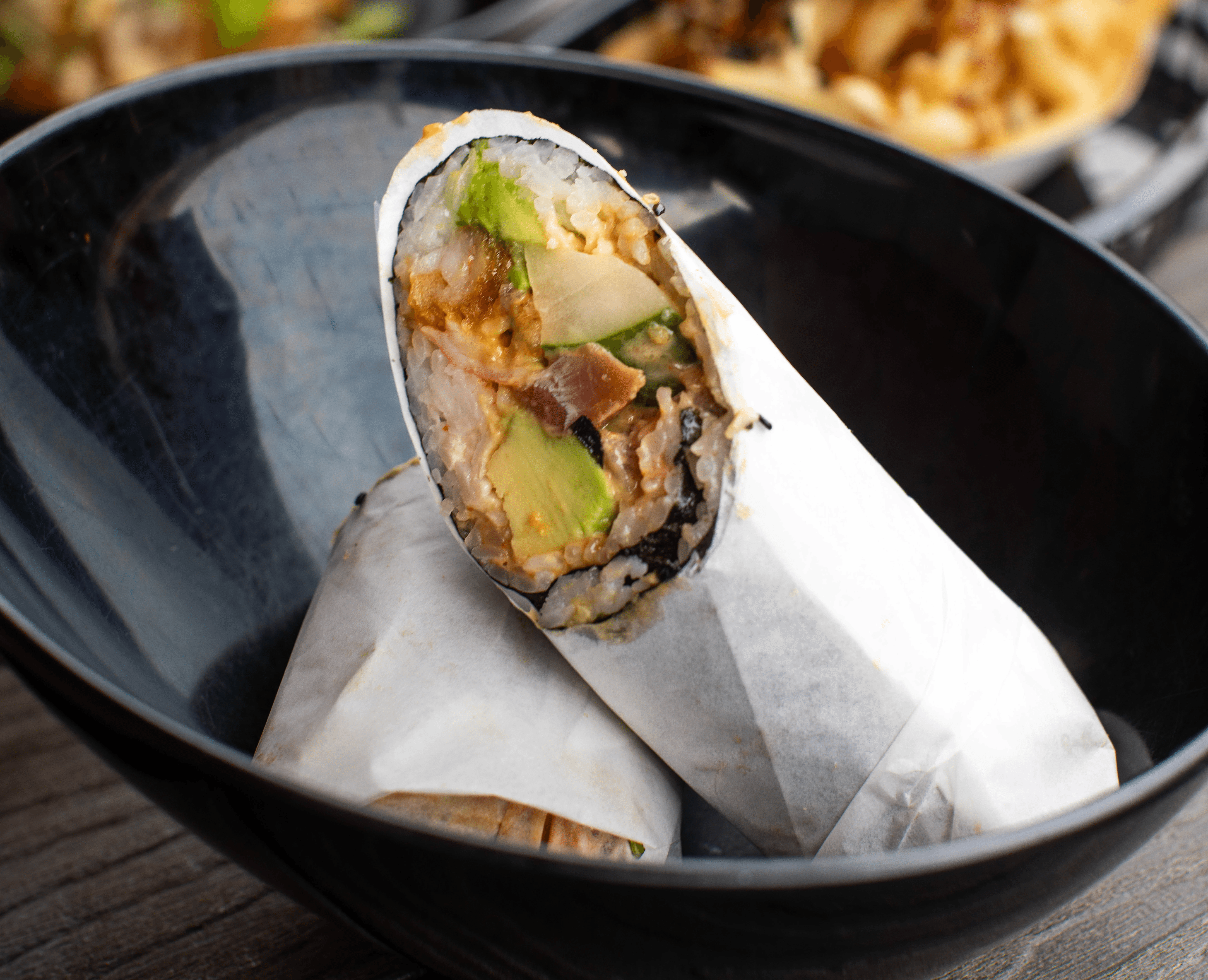 A thick cut of steak with prominent grill marks sizzles over hot coals and flames on a barbecue, with blurred figures of people and drinks in the background, suggesting an outdoor gathering.