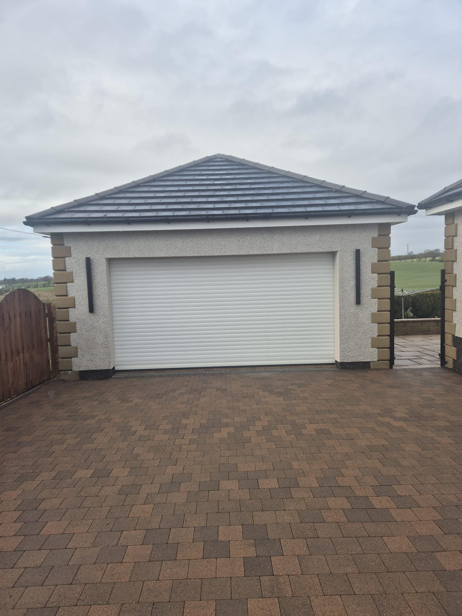 A modern single-car garage with a white roller door and small rectangular windows on each side. A brick driveway leads to the garage, with a cloudy sky above.