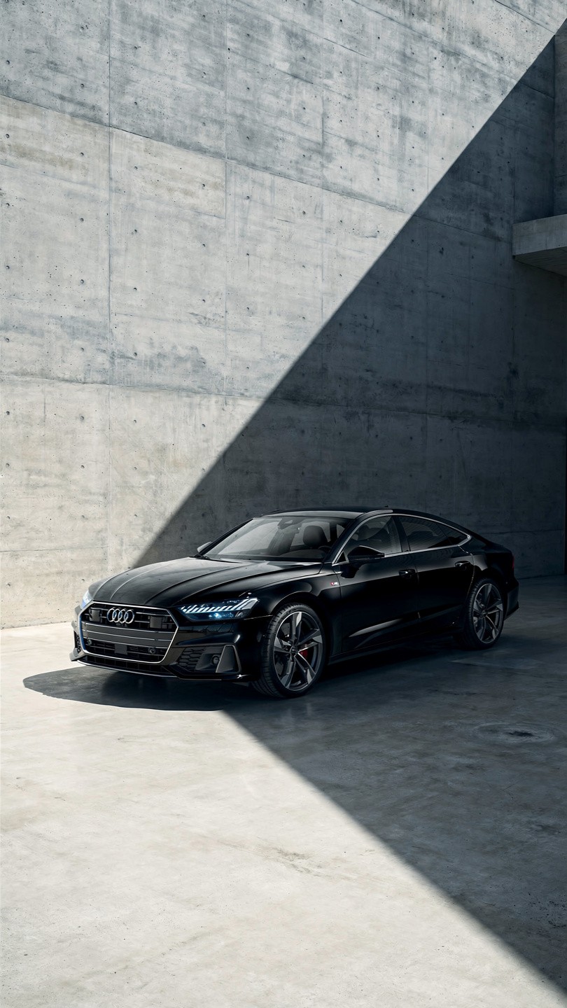 Sleek black car in a modern concrete space, partially lit by a diagonal beam of sunlight, creating a dramatic contrast. The scene feels sophisticated and elegant.
