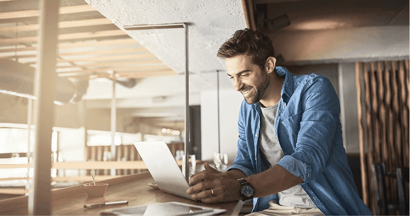 Smiling man working on a laptop in a modern café, wearing a casual denim shirt and enjoying remote work.