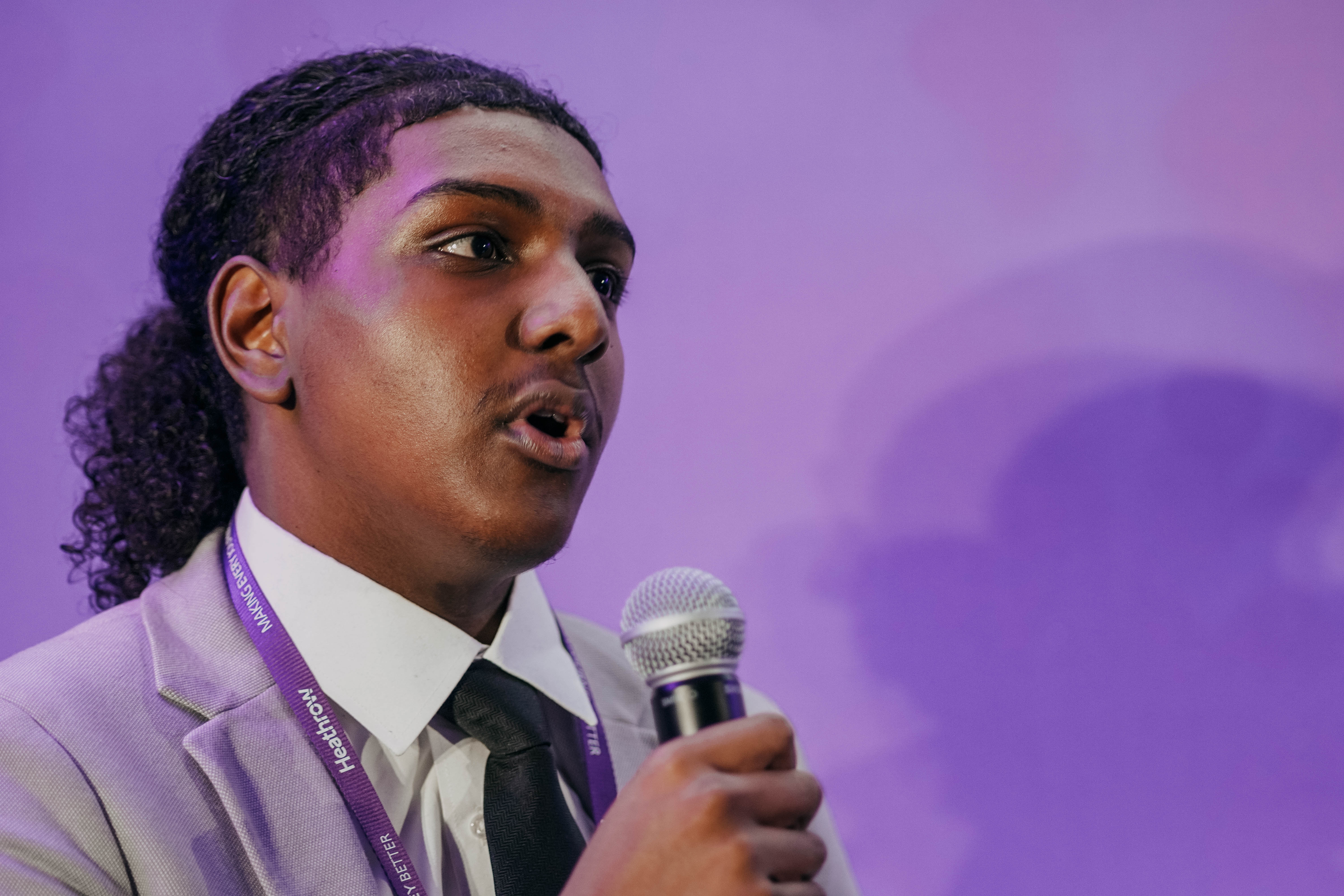 A young man with his hair tied back speaks intently into a microphone against a purple background, wearing a suit jacket and a Heathrow lanyard.