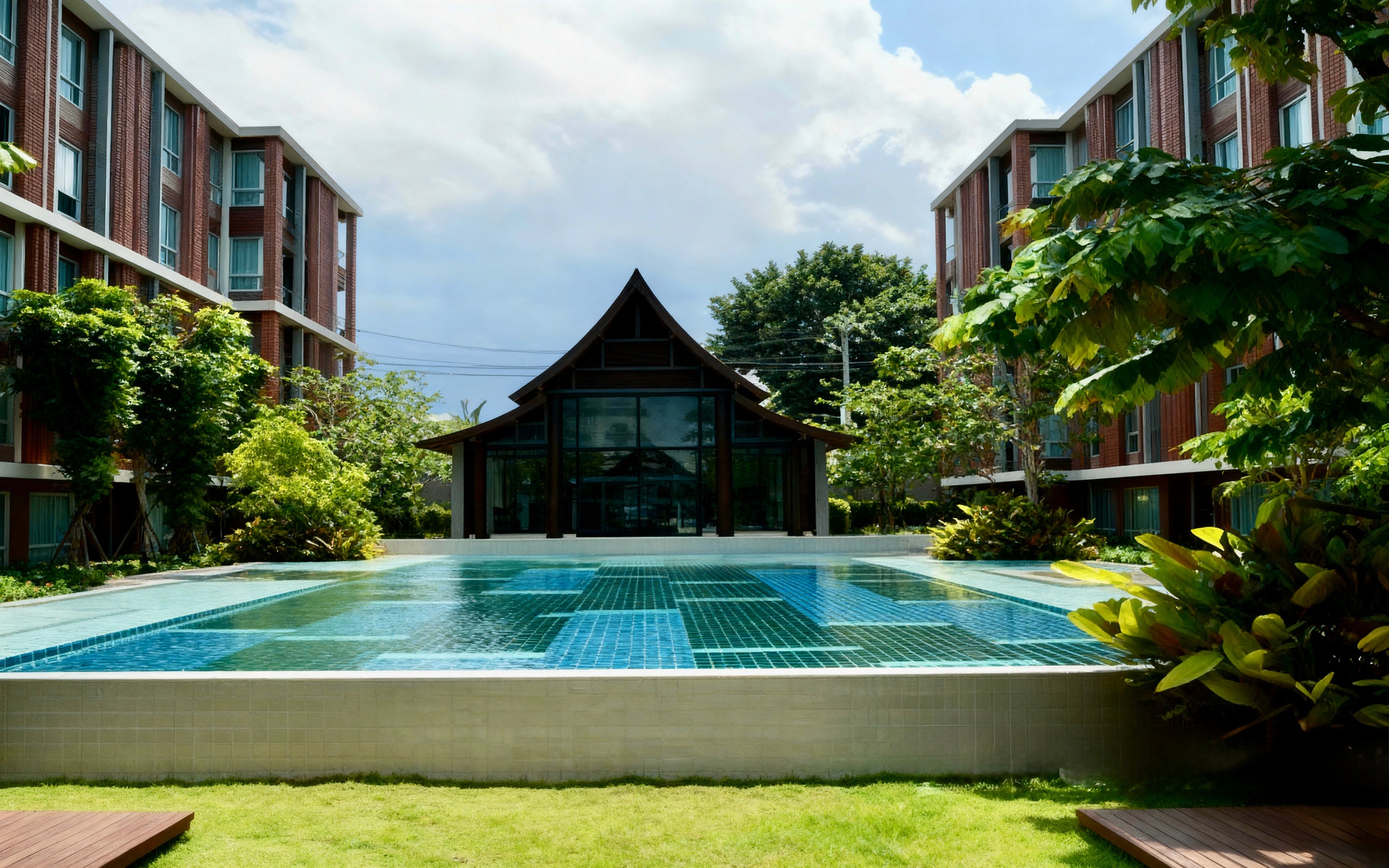 Modern apartment complex with a central swimming pool surrounded by tropical greenery and red-brick buildings under a bright blue sky in Santitham, Chiang Mai.