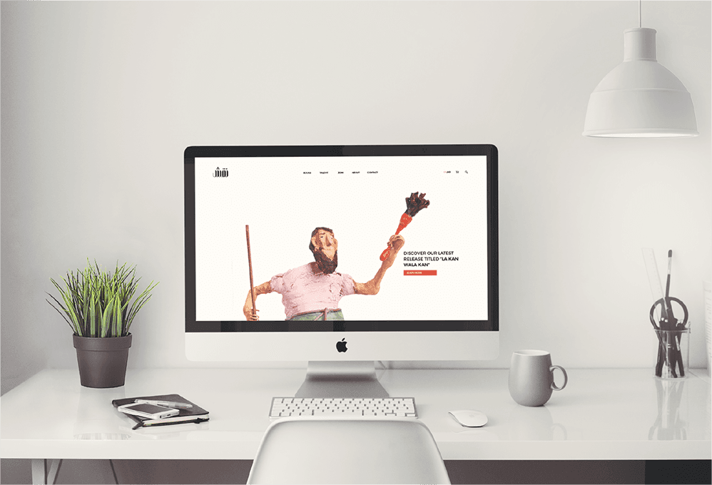 An Apple iMac computer displays a webpage with a man holding a broomstick. The webpage’s text reads “OUR OURLY LATEST RELEASE TITLED ‘LA KAN WALA’”. The computer sits on a white desk next to a potted plant, a notebook, a smartphone, a mug, and a pair of scissors. A white pendant light hangs above the desk.