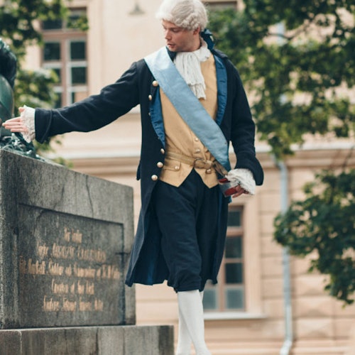 Mies, pukeutuneena historiallisesti siniseen nauhaan ja tricorne-hattuun, seisoo kivijalan vieressä. Taustalla on puita ja rakennus.