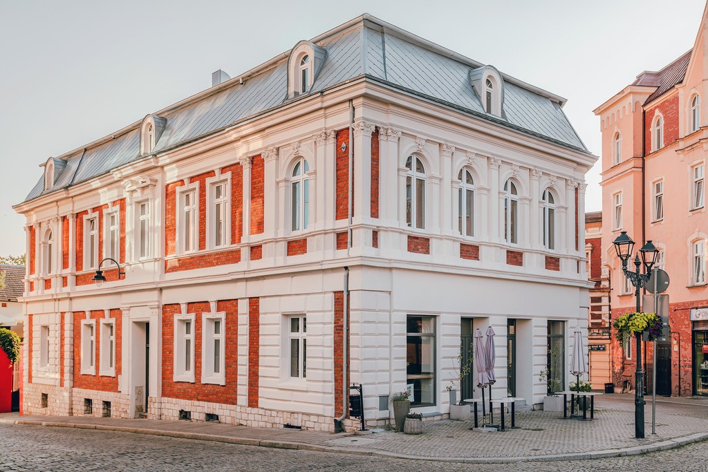 Piękny budynek w stylu klasycystycznym z czerwonymi cegłami. Na rogu ulicy z białymi elementami. Stoliki i parasole na zewnątrz.