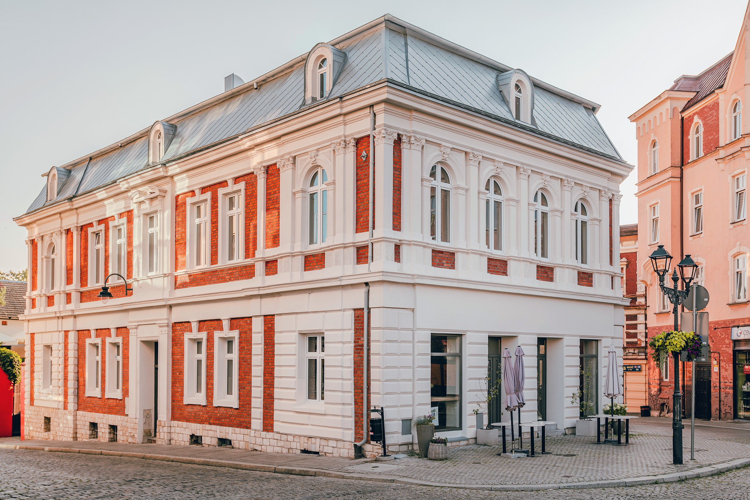 Piękny budynek w stylu klasycystycznym z czerwonymi cegłami. Na rogu ulicy z białymi elementami. Stoliki i parasole na zewnątrz.