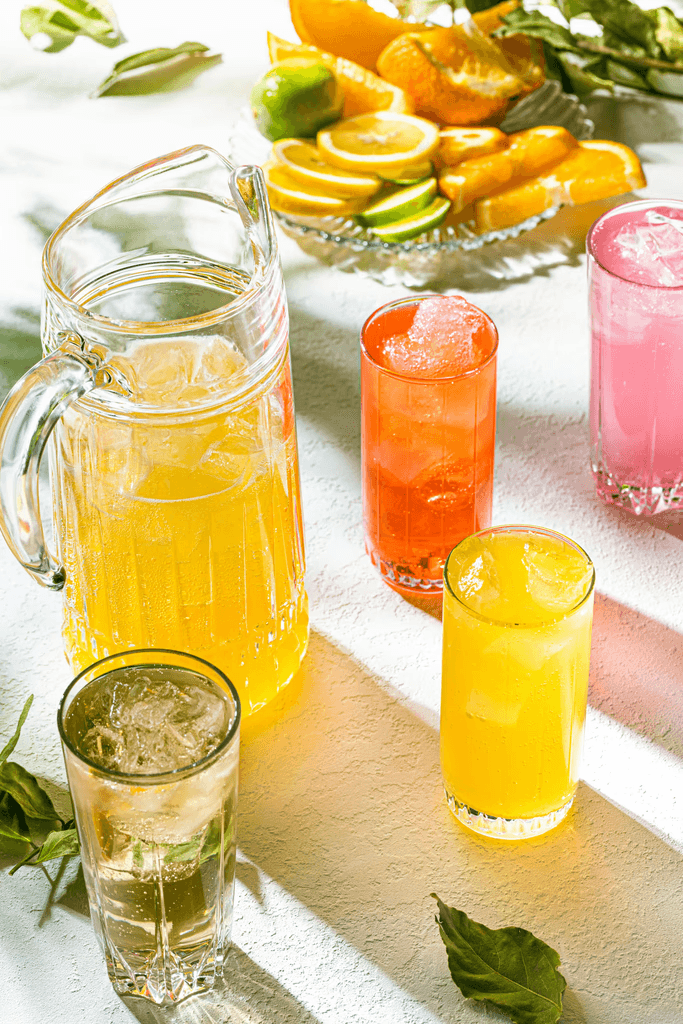 Brightly lit scene with a pitcher and glasses filled with colorful citrus drinks, surrounded by slices of lemon, lime, and orange on a textured white surface.
