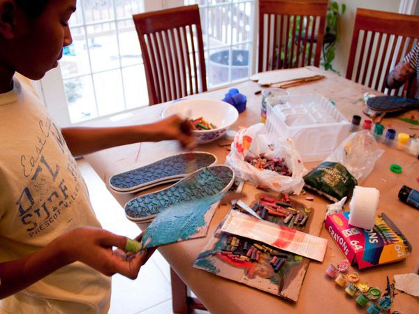 A child wearing a light-colored shirt with blue lettering is painting a pair of blue-gray patterned shoes on a light brown table. The child is using a paintbrush in their right hand and holding a piece of blue-green paint in their left hand. A white bowl containing colorful items sits on the table near the child, along with several small containers of paint and crayons. A plastic bag with colorful items is also on the table. The table is positioned near a window with white trim and wooden chairs are visible in the background.
