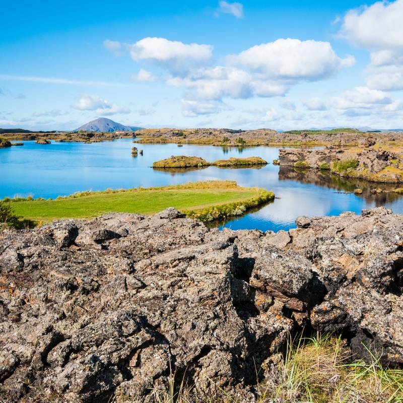 Volcanic lava formations surround the tranquil waters of Lake Mývatn in North Iceland, showcasing the dramatic landscape created by thousands of years of volcanic activity and lava flows.