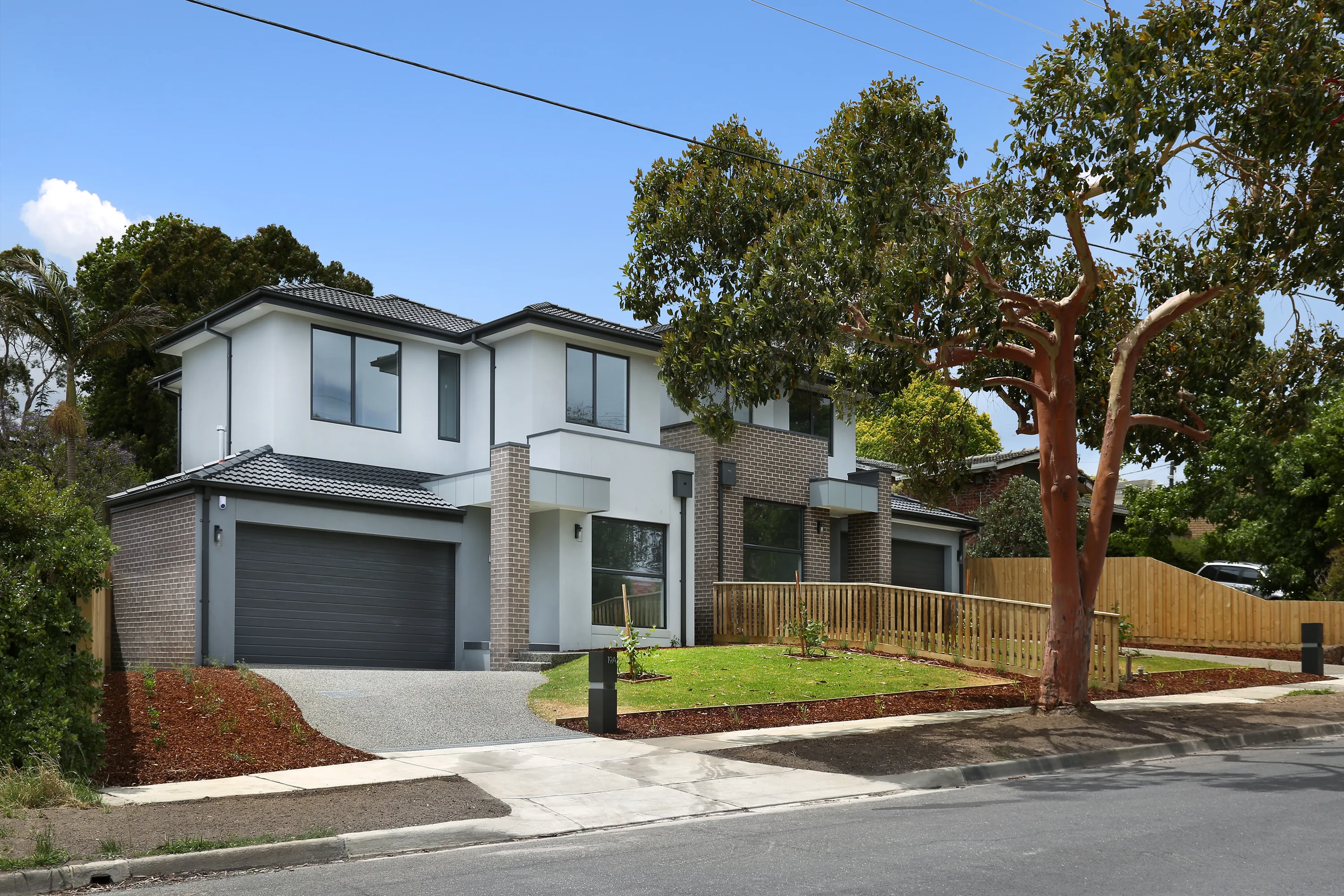 Street view of the two Waverley homes, contemporary two-storey residences in white render and dark brick with grey tiled roofs, charcoal garage doors and landscaped front gardens among gum trees