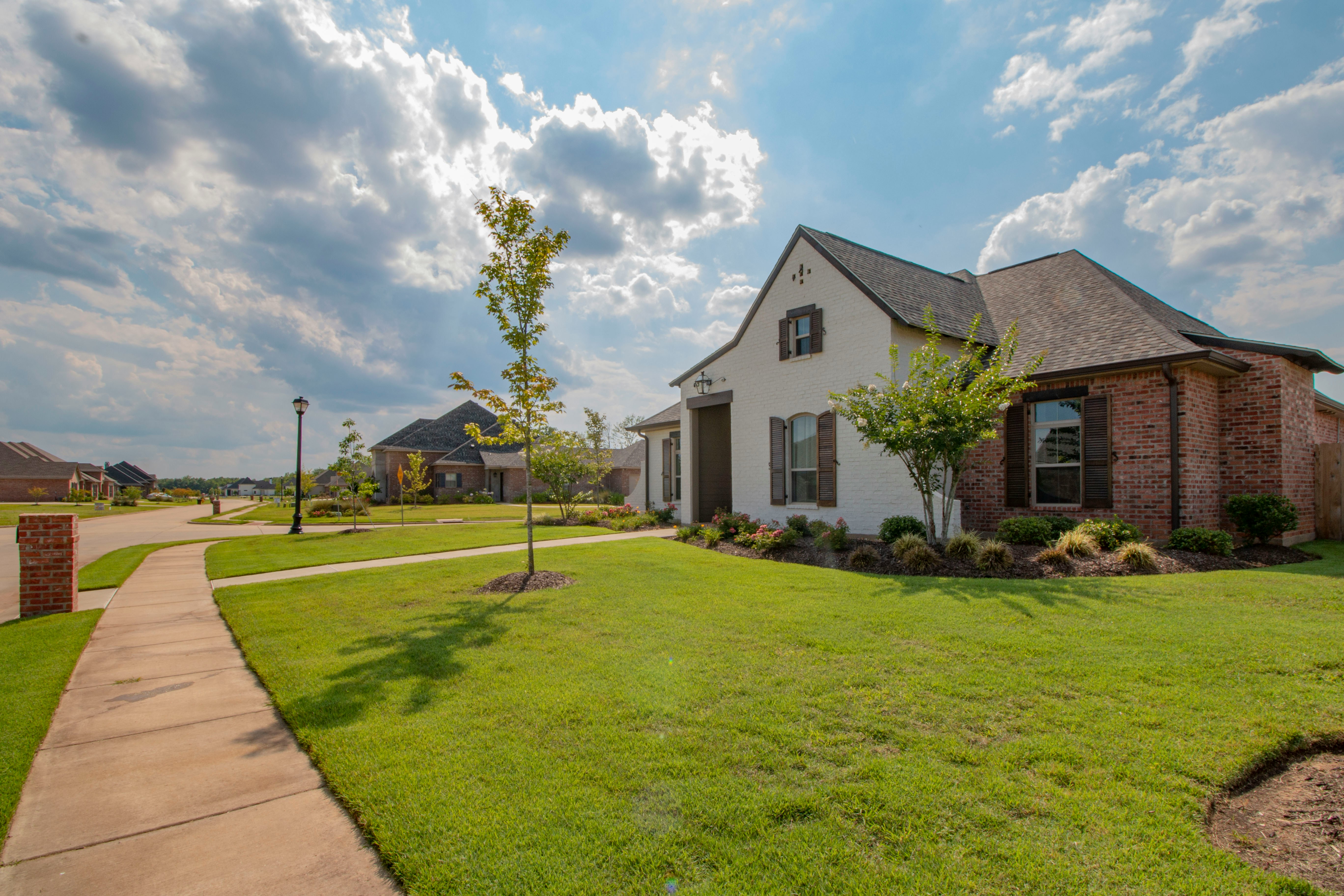 Single-family suburban house with a mix of brick and white exterior, gabled roof, front lawn, and landscaped yard under a partly cloudy sky.