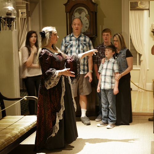 在一个有古董钟、长椅和柔和灯光的房间里,一位穿着古装的女士与一个站着的家庭交谈。