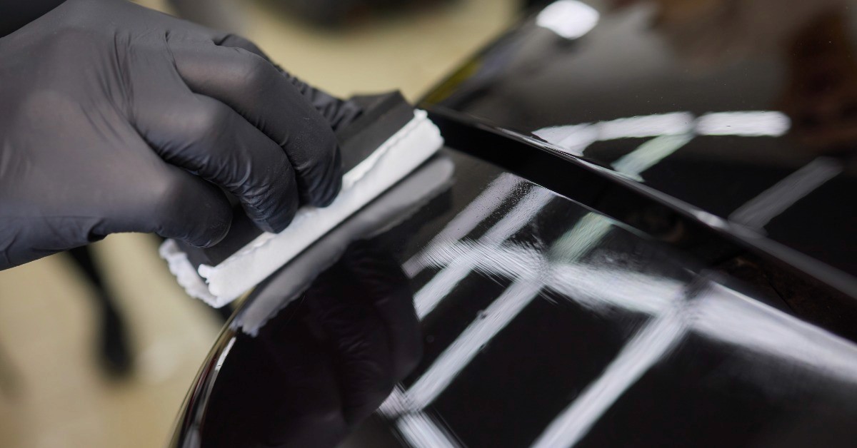 A black-gloved hand is pressing a white cloth against a glossy black car panel. Bright lights reflect onto the vehicle.