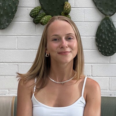 A woman smiling while seated in front of a white brick wall decorated with large cactus leaves.