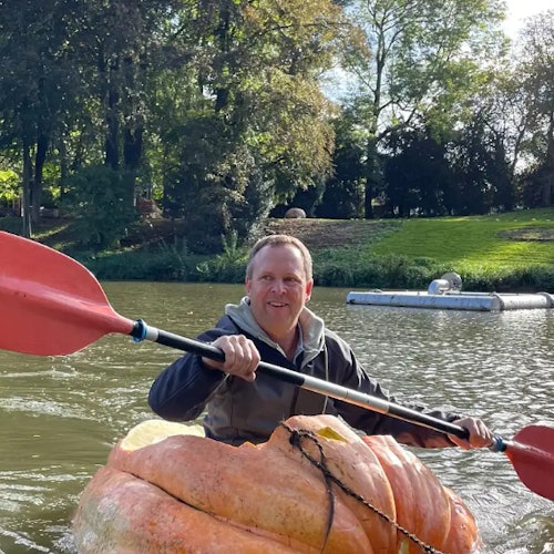 A man paddles a pumpkin boat with red oars on a calm lake, surrounded by lush trees and greenery.