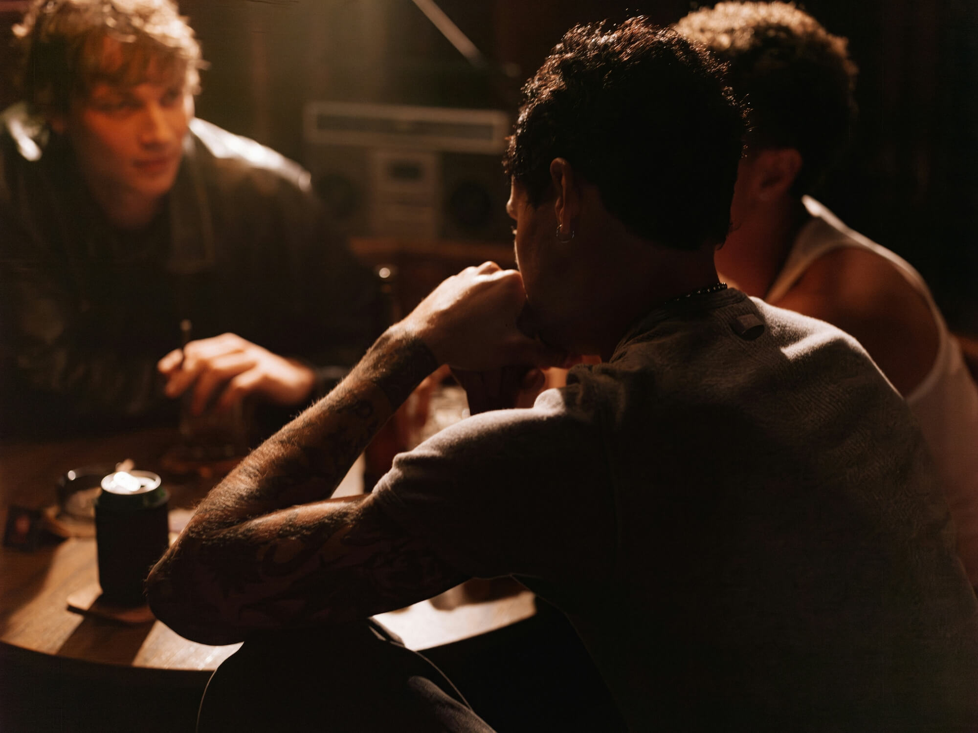 Three people sit around a dimly lit table with drinks and cards, conveying a relaxed, intimate atmosphere. A boombox is visible in the background.