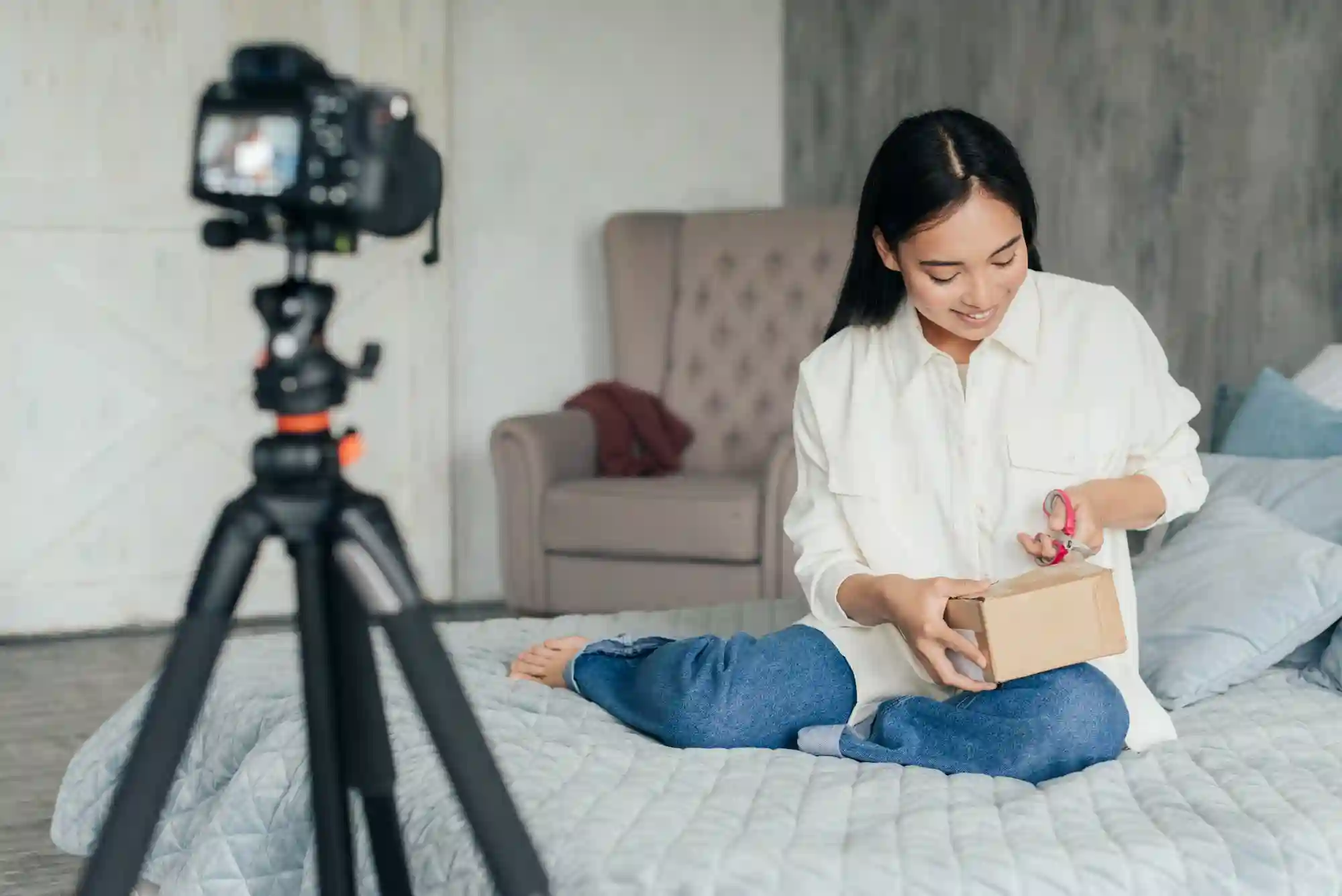 Asian woman recording an unboxing video of a package while sitting on a bed for her vlog.
