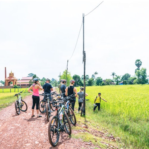 A group of cyclists stands with their bikes on a dirt path next to a green field, with a building visible in the background.