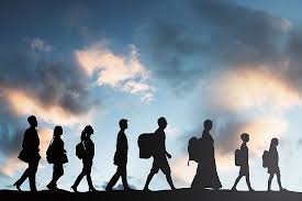 A horizontal silhouette of a diverse group of people, including adults and children, walking in a single file line against a dramatic sunset or twilight sky filled with blue, orange, and dark clouds. Many of the figures appear to be carrying bags or backpacks.