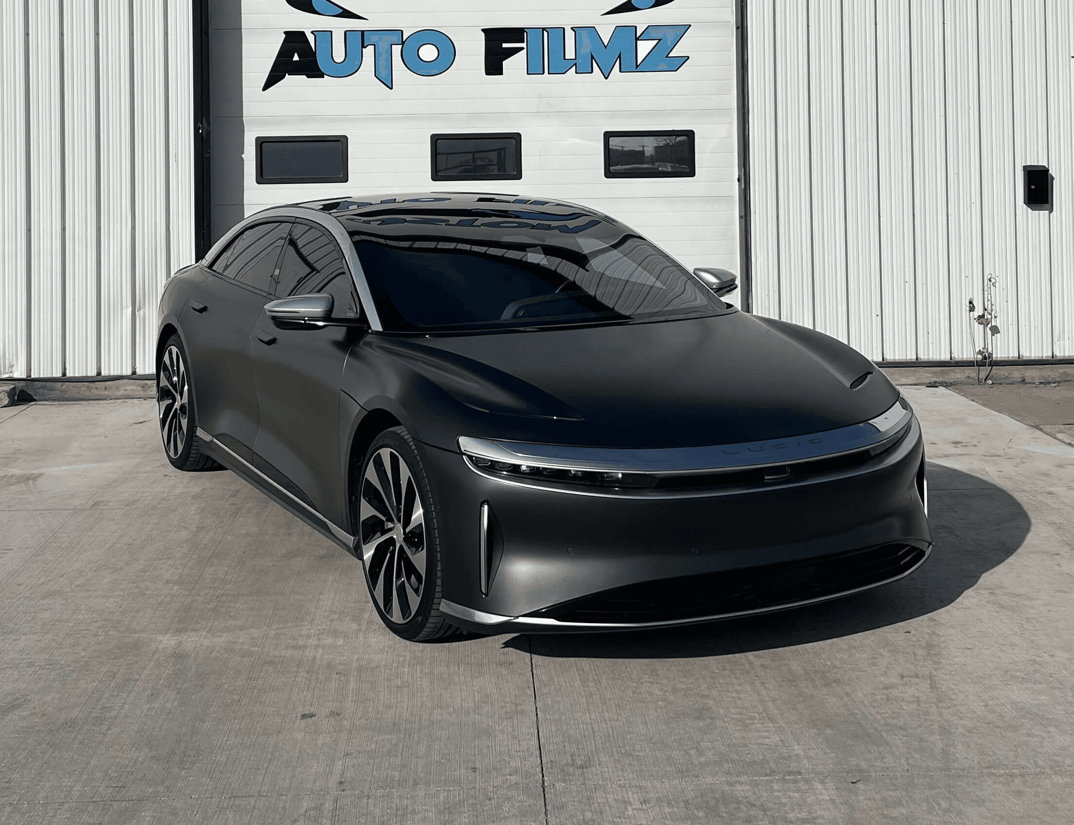 A matte gray Lucid Air luxury electric sedan is parked on a concrete driveway in front of a white industrial building.